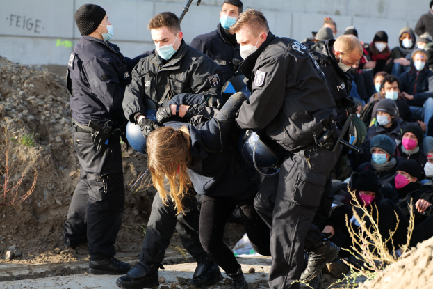 Eine Gruppe maskierter Personen in Mützen, einige bewaffnet, umringt eine Frau, die von der Polizei eskortiert wird, mit Pflanzen, Steinen und einer Wand mit Protesttext im Hintergrund.
