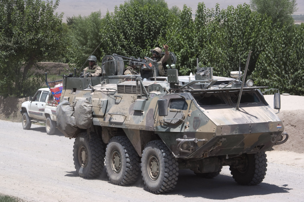 Gepanzerter Personentransporter und Lkw fahren auf einer Schotterstraße mit bewaffneten Individuen in Helmen, umgeben von Bäumen, Hügeln und einem klaren Himmel.
