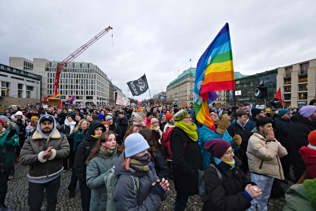 Eine große Gruppe von Menschen mit Fahnen und Schildern, die vor einem Gebäude während einer LGBTQ+-Rechtsdemo in Berlin stehen, mit Gebäuden, einem Kran und einem bewölkten Himmel im Hintergrund.