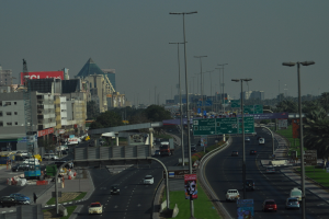 Eine Luftaufnahme einer Stadt mit Gebäuden, Türmen, Fahrzeugen, Werbetafeln, Schildern, Straßen, Bäumen, Straßenlaternen, Menschen, Verkehrskegeln und Informationsanzeigen unter einem klaren Himmel.