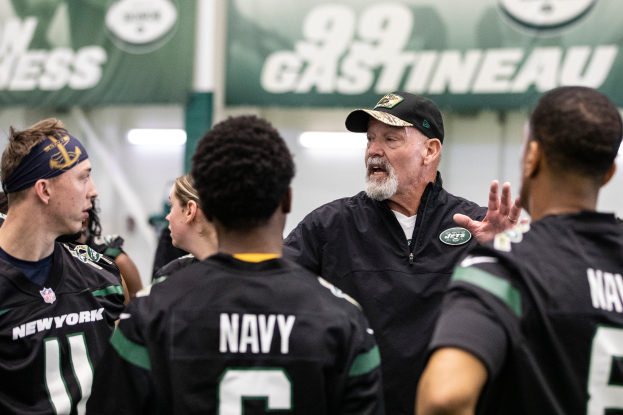 Eine Gruppe von Männern, die zusammen auf einem Feld stehen, mit einem Mann in der Mitte, der eine Mütze trägt, wahrscheinlich Jets-Headcoach Mike McCarthy, der mit seinem Team spricht, mit Bannern und Lichtern im Hintergrund, die auf ein Stadion oder eine Sportanlage hindeuten.