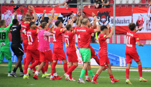 Gruppe von Fußballspielern, die ein Tor feiern, mit Flaschen haltenden und klatschenden Spielern, Bannern, Zaun, Stühlen und einem Dach mit Beleuchtung im Hintergrund.