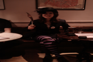 Eine Frau sitzt auf einer Bank und hält ein Messer in der Hand, mit einem Tisch voller Gegenstände vor ihr und einer anderen Person neben ihr.