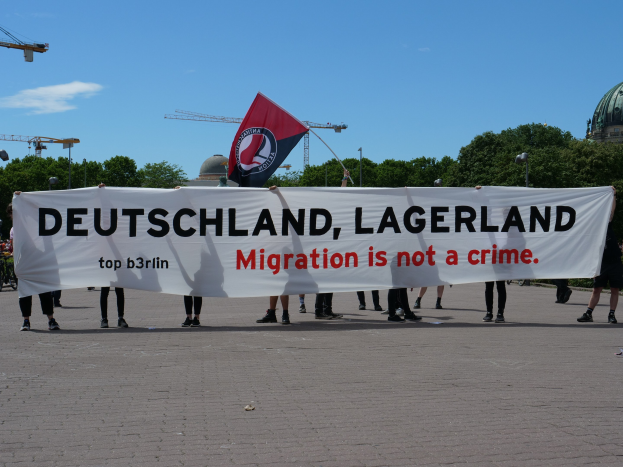 Eine Gruppe von Menschen hält ein Transparent mit der Aufschrift 'Deutschland, Lagerland Migration ist kein Verbrechen' und eine Flagge, im Hintergrund sind Straßenlaternen, Straßen, Bäume, Kräne, ein Gebäude und ein bewölkter Himmel zu sehen.