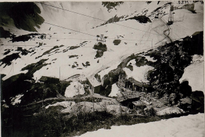 Ein Schwarz-Weiß-Foto eines Skilifts, der einen schneebedeckten Berg hinauffährt, mit Häusern, Pfählen und Drähten im Vordergrund und Text am unteren Rand.