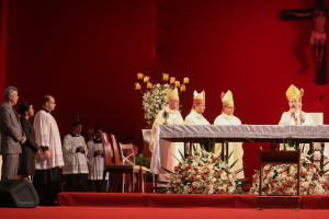 Vier Personen, die stehen, einige halten Kerzen auf Ständern, eine Person hält ein Mikrofon, eine Gruppe von Personen steht, Stühle, ein Lautsprecher, Blumen und heilige Kreuze an der Wand im Hintergrund.