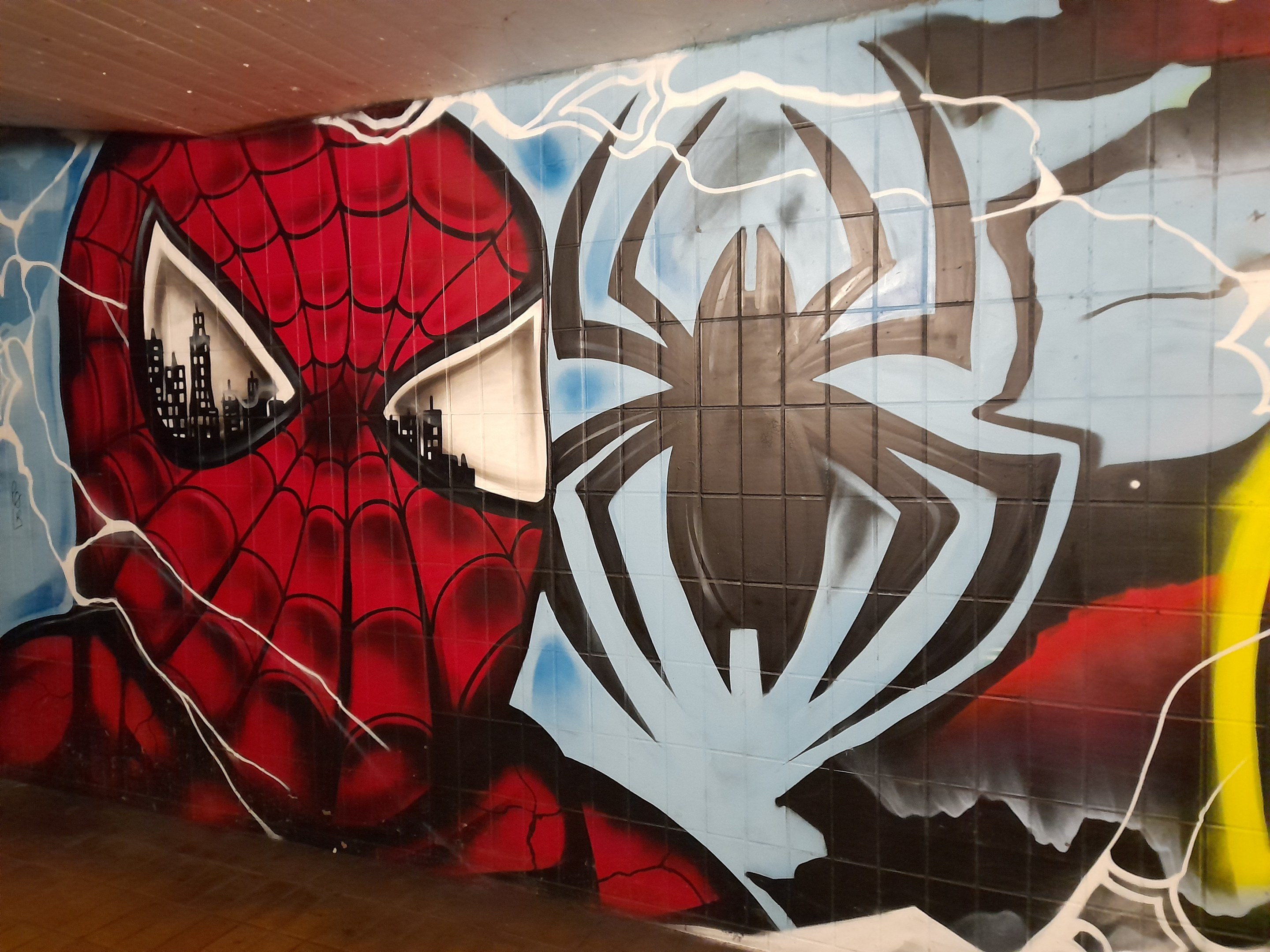 Ein buntes Wandgemälde von Spider-Man in rot und blau mit einem Webmuster und einem großen Spinnenlogo, gemalt an der Seite eines Gebäudes.