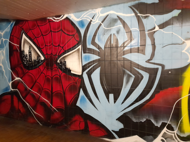 Ein buntes Wandgemälde von Spider-Man in rot und blau mit einem Webmuster und einem großen Spinnenlogo, gemalt an der Seite eines Gebäudes.