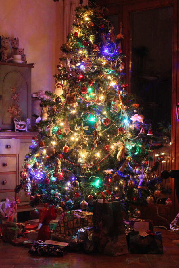 Ein Weihnachtsbaum mit Lichtern und Dekoration, Geschenke auf dem Boden, ein Schrank mit Gegenständen links und ein Fenster an der rechten Wand.