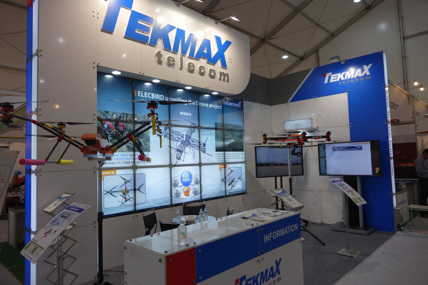 Ein Messestand für Tekmax Telecom mit einer großen Wandanzeige, einem Tisch mit Produkten, zwei Fernsehern, Informationsschildern, Deckenleuchten und einigen Personen.