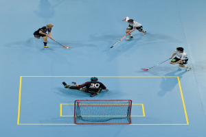 Drei Personen in Sportkleidung spielen Hockey, wobei eine Person rechts einen Ball mit einem Hockeystock schlägt und eine Person hinter einem Netz auf dem Boden sitzt.