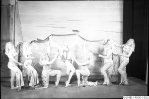 Eine Gruppe von Frauen in Bikinis tanzt auf einer Bühne mit einem Netz im Hintergrund und Vorhängen, die die Szene einrahmen, dargestellt in Schwarz-Weiß.