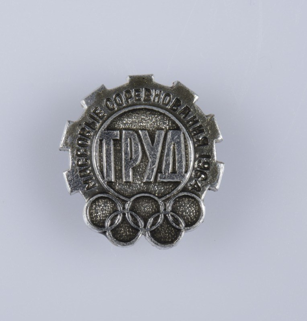 Eine silberne Anstecknadel mit der Aufschrift 'Olympische Spiele' auf einem weißen Hintergrund.