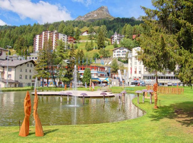 Ein zentraler Wasserplatz mit Fontänen, umgeben von Gebäuden, Bäumen und Bergen, mit Gras am unteren Bildrand.