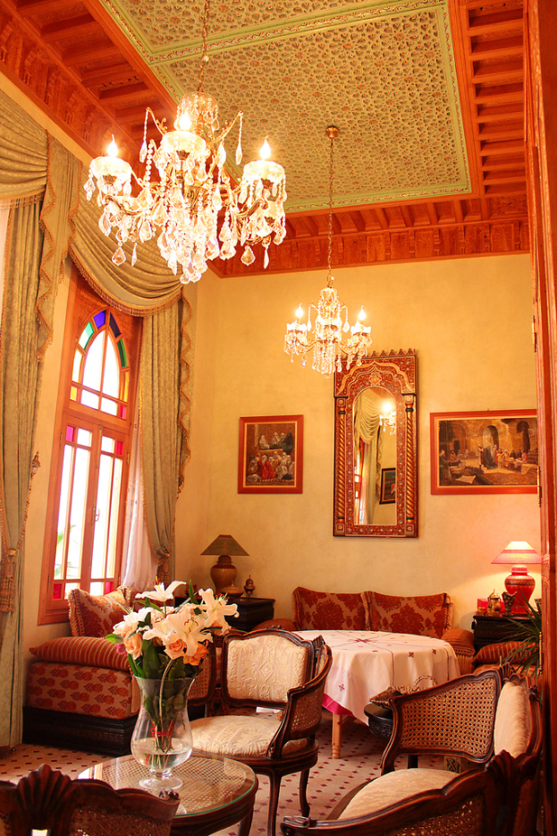 Ein Wohnzimmer mit einem Tisch und Blumentopf im Vordergrund, umgeben von Stühlen, Sofas und einem Tisch mit weißer Decke im Hintergrund, gerahmte Bilder an der Wand, von der Decke hängende Lampen und Vorhänge mit einem Fenster links.