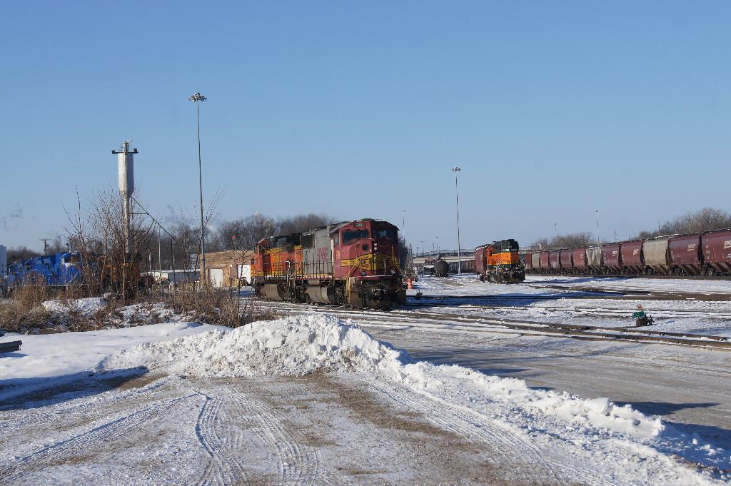Eine Lokomotive ist vorne positioniert mit einem Waggon auf der rechten Seite, geparkt auf Schienen mit Schnee auf dem Boden; trockene Bäume und ein Strommast sind im Hintergrund sichtbar.