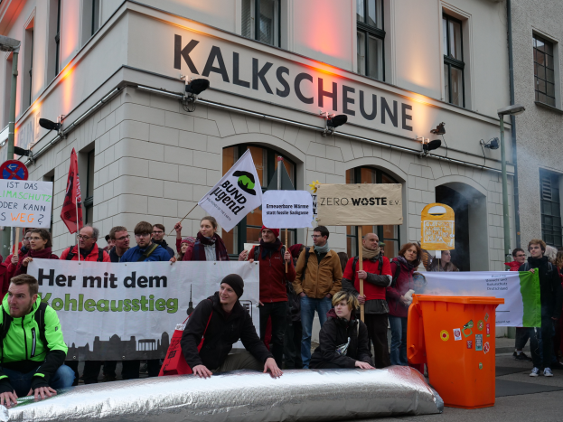 Eine Gruppe von Menschen steht vor einem Gebäude und hält Schilder und Plakate hoch, während im Vordergrund zwei Personen auf einem Gegenstand sitzen und ein Müllcontainer rechts zu sehen ist, bei einer Demonstration in Deutschland.